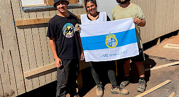 Voluntarios y habitantes damnificados frente a una vivienda sosteniendo bandera "La UC sirve a Chile"