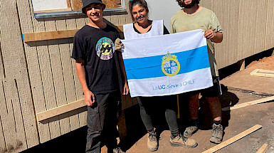 Voluntarios y habitantes damnificados frente a una vivienda sosteniendo bandera "La UC sirve a Chile"