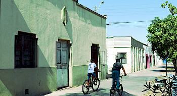 Niños andan en bicicleta por una vereda y se aprecia el frontis de una casa antigua.