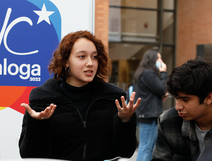 Mujer joven hablando y elevando sus manos, junto a un joven a su lado.