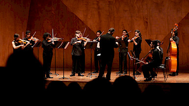 Orchestra playing on a stage with audience silhouettes.