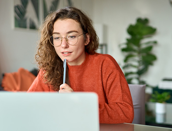 Una mujer observa la pantalla de un computador sosteniendo un lápiz y usando un sweater color naranja.