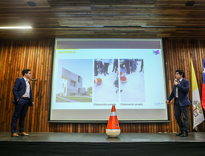 Dos jóvenes presentando, con un cono de seguridad naranjo al medio del escenario