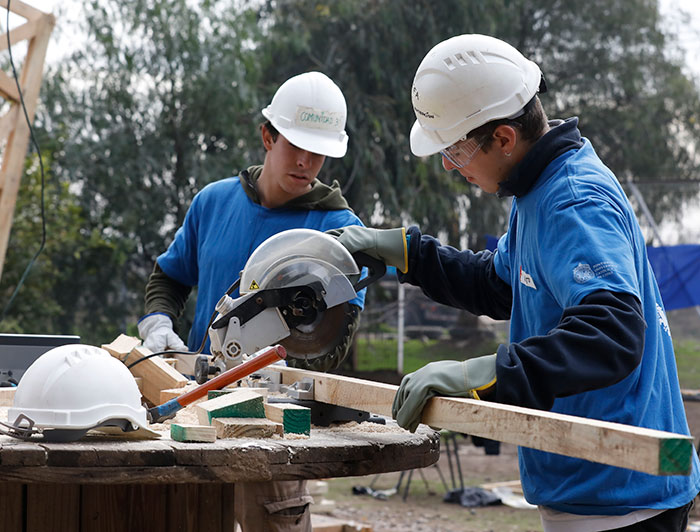 Dos jóvenes con polera azul y casco blanco cortan una madera 
