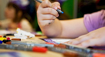 Mano de una niña sosteniendo un lápiz de color y un cuaderno, junto a lápices dispersos en una mesa.