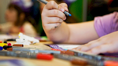 Mano de una niña sosteniendo un lápiz de color y un cuaderno, junto a lápices dispersos en una mesa.