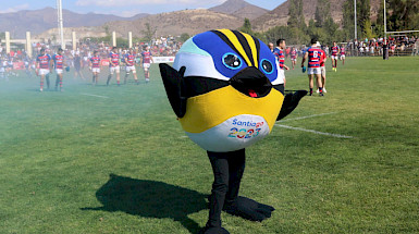 Mascota Fiu de los XIX Juegos Panamericanos y VII Parapanamericanos Santiago 2023, en una cancha con jugadores de rugby en el fondo.