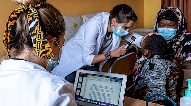 One doctor looks at her computer, while another checks on a child who is in the arms of his mother.
