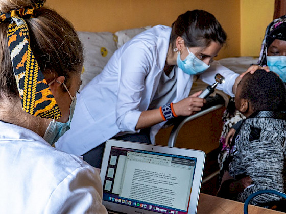 One doctor looks at her computer, while another checks on a child who is in the arms of his mother.