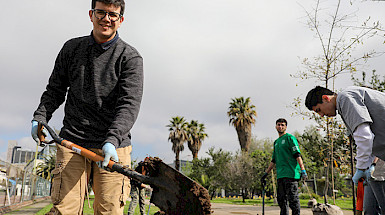 Estudiante sostiene una pala mientras participa de la forestación del campus San Joaquín, aparece un par de estudiantes más al fondo.
