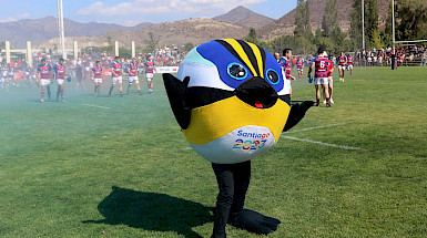 Image of the official mascot of the Games, Fiu, a seven-colored bird. He is smiling and with his arms raised in the middle of a field with rugby players behind him.