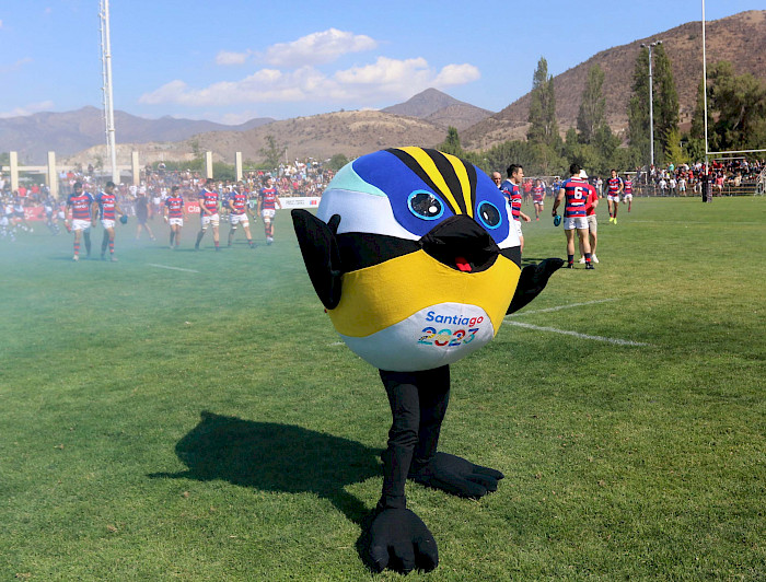 Image of the official mascot of the Games, Fiu, a seven-colored bird. He is smiling and with his arms raised in the middle of a field with rugby players behind him.