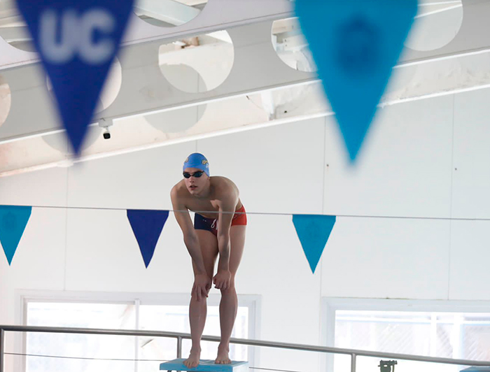 Student Elías Ardiles wearing a swimsuit and a cap between blue flags, one with the "UC" logo, ready to jump into an Olympic pool. He rests his hands on his knees as he looks concetrated.