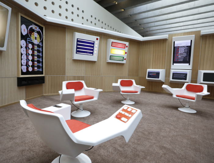 Cybersyn room, it is similar to a spaceship. It is illuminated, has wooden walls, screens, lights and white and red seats.