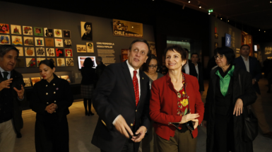 UC Chile President Ignacio Sánchez with Interior Minister Carolina Tohá looking at the exhibition, which has paintings alluding to the time. The room is dimly lit and there are more people walking around.