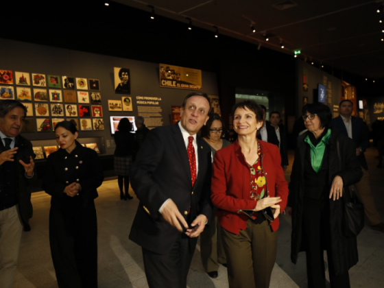 UC Chile President Ignacio Sánchez with Interior Minister Carolina Tohá looking at the exhibition, which has paintings alluding to the time. The room is dimly lit and there are more people walking around.