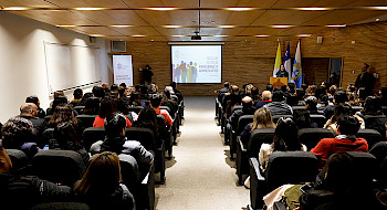 Ceremonia de lanzamiento nueva política para administrativos y profesionales UC, donde se ve el público sentado en un auditorio con la presentación al fondo y la directora de Personas en el podio.
