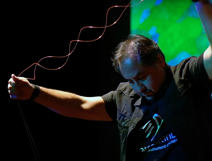 El profesor Rodrigo Cádiz durante su concierto Sibila de 2022, sosteniendo unos cables, con fondo negro y luces verdes y azules. 