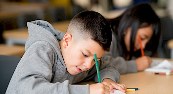 Niño en primer plano escribe con un lápiz sobre un cuaderno en un escritorio, junto a una niña.