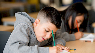 Niño en primer plano escribe con un lápiz sobre un cuaderno en un escritorio, junto a una niña.