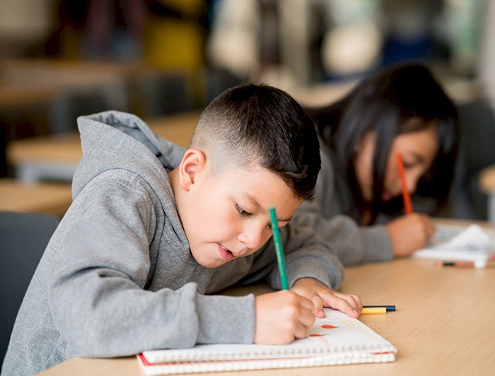 Niño en primer plano escribe con un lápiz sobre un cuaderno en un escritorio, junto a una niña.