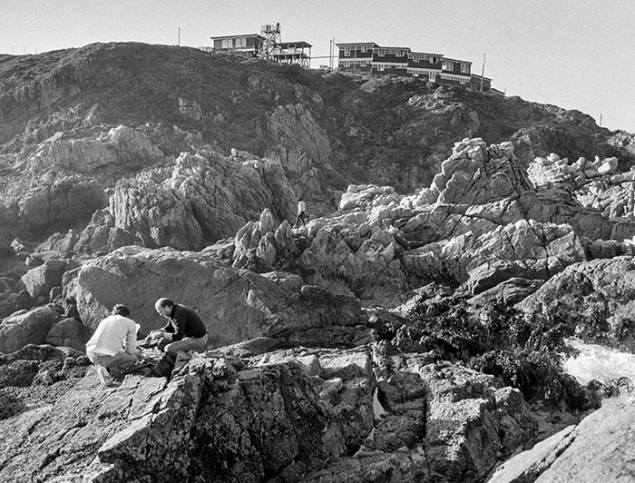 Black and white image showing two people investigating the rocks and the original station above.