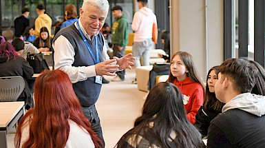 El director del programa Bachillerato Inicia conversando con un grupo de postulantes del programa en una sala; se ven otros jóvenes al fondo.