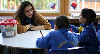 mujer sentada en una mesa escolar frente a dos niños