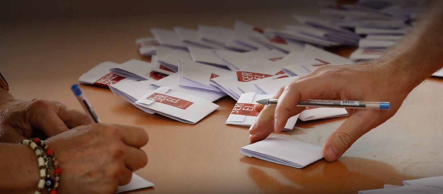 Manos sosteniendo o escribiendo sobre un voto, sobre una mesa en que se ven votos cerrados de fondo.