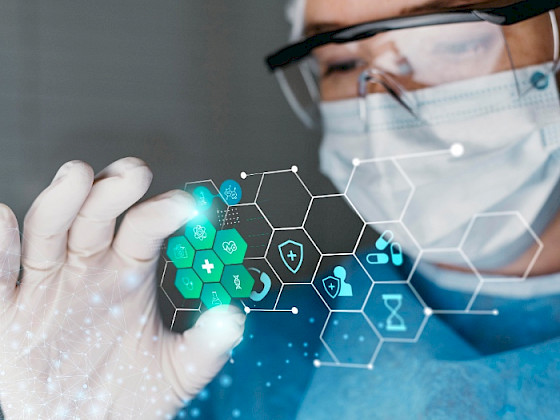 Persona usando mascarilla, lentes, bata celeste y guantes, interactúa con una pantalla virtual
