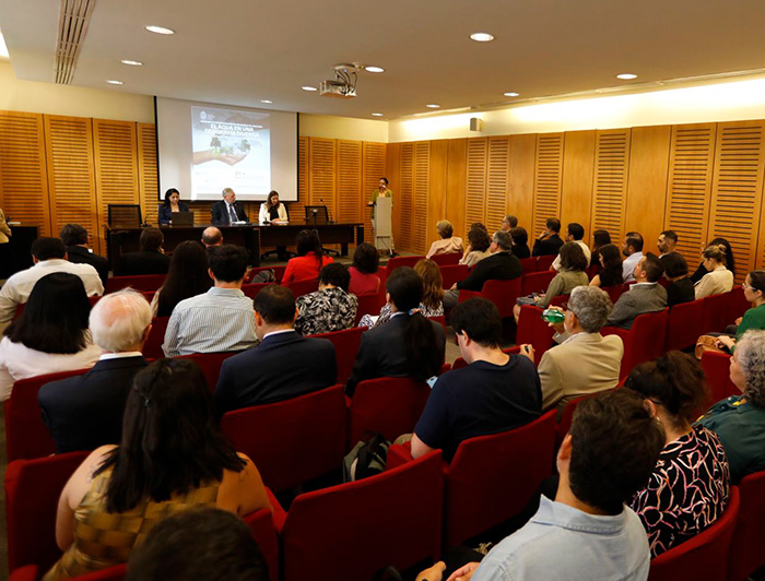 Público en una sala frente a tres expositores sentados en una testera y una imagen proyectada al fondo.