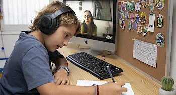 Niño escribe sobre un cuaderno usando audífonnos y junto a un computador donde se ve una mujer.