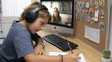 Niño escribe sobre un cuaderno usando audífonnos y junto a un computador donde se ve una mujer.