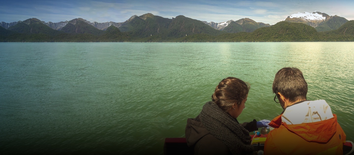 Dos jóvenes toman muestras en el agua con montañas de fondo.