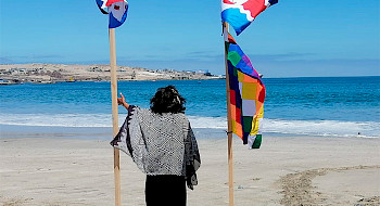 Mujer de espalda mira el mar usando vestimenta de la cultura Chango junto a banderas de pueblos originarios.