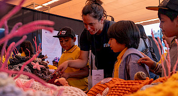 La investigadora de Núcleo NUTME, Celeste Kroeger, le muestra a unos niños la exposición Tramares, Tejiendo Arrecifes.