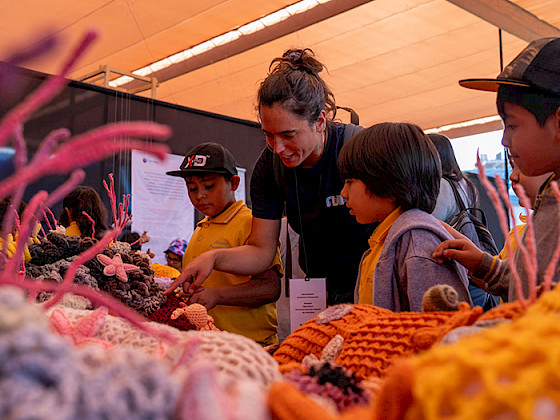 La investigadora de Núcleo NUTME, Celeste Kroeger, le muestra a unos niños la exposición Tramares, Tejiendo Arrecifes.