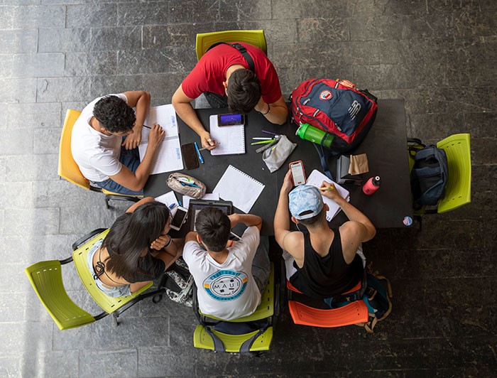 Vista desde arriba de un grupo de jóvenes sentados en una mesa estudiando, con sus cuadernos, teléfonos y computador.