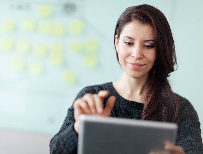 Mujer sostiene una tablet y de fondo se divisan post it amarillos pegados en una pared.