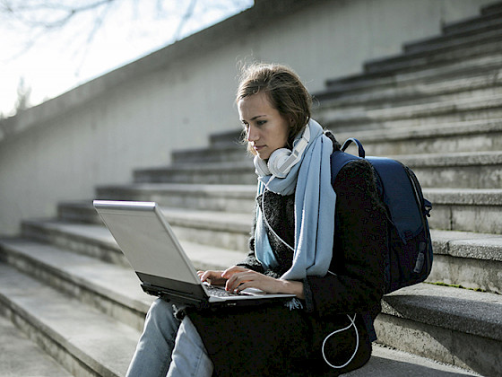 Mujer joven sentada en una escalera trabaja en su notebook, lleva unos audífonos y una mochila.