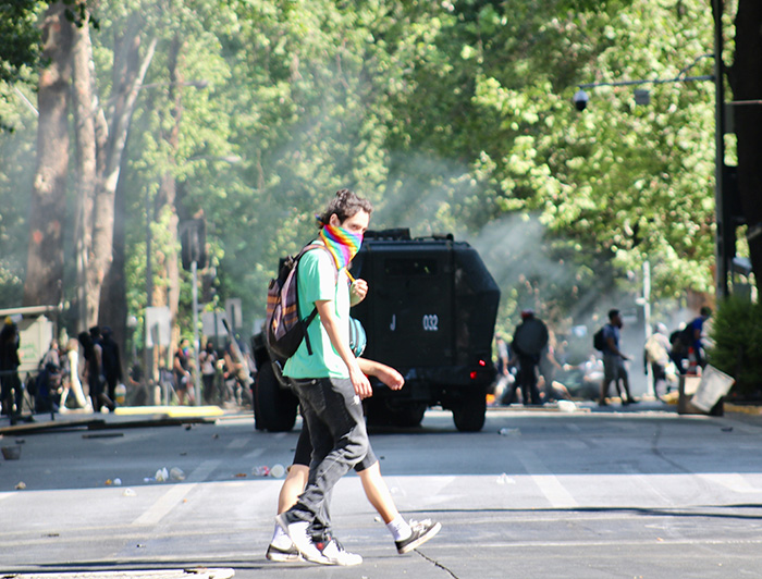 Joven con la cara cubierta con un pañuelo camina por la calle, de fondo un carro policial y una manifestación. 