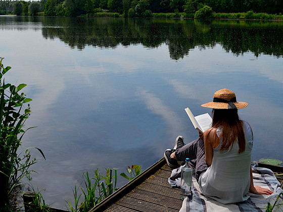 Mujer leyendo en un muelle con agua y vegetación de fondo