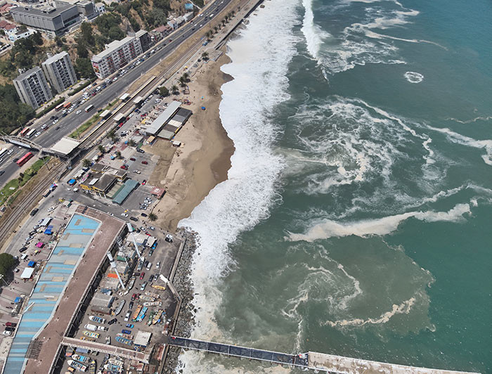 Drone image of powerful sea swells hitting the Chilean coast, with nearby buildings and infrastructure highlighting the risk of beach loss due to intensified wave activity and coastal urbanization.