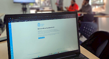 Computador que muestra la plataforma de AyudantIA; se ven profesores en capacitación al fondo de la sala.