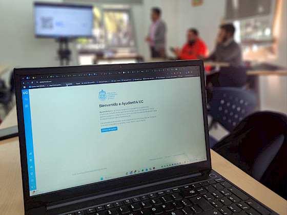 Computador que muestra la plataforma de AyudantIA; se ven profesores en capacitación al fondo de la sala.