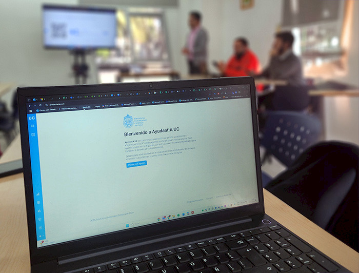 Computador que muestra la plataforma de AyudantIA; se ven profesores en capacitación al fondo de la sala.