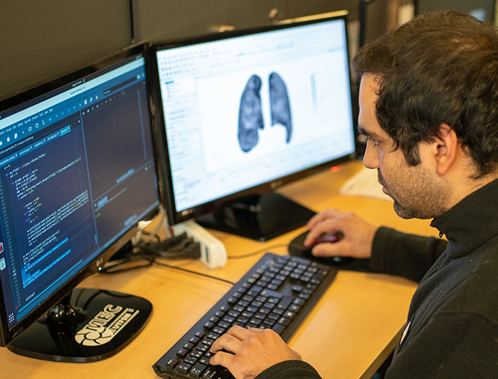 Investigador trabajando en un computador con dos pantalles, en las que se muestra un código y la imagen de un pulmón.