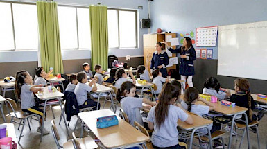 Dos profesoras con delantal azul imparten clases a niños y niñas en sala de clases.