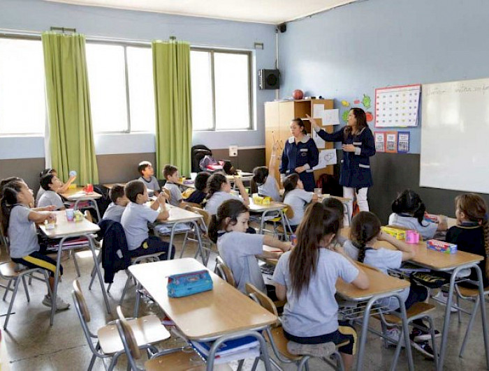 Dos profesoras con delantal azul imparten clases a niños y niñas en sala de clases.