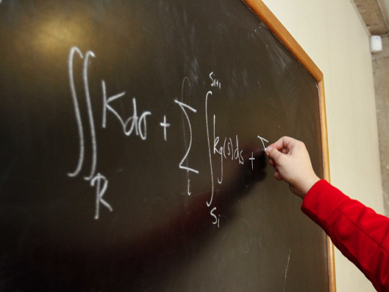 Mano escribiendo con tiza una fórmula matemática sobre una pizarra negra.